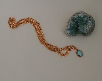 Könnte beinhalten: Eine goldfarbene Halskette mit einem hellblauen, ovalen Anhänger. Die Kette liegt auf einer weißen Oberfläche neben einem rauen, türkisgrünen Stein.