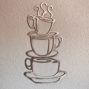 May include: Metal wall art featuring a stack of three coffee cups with steam rising from the top cup. The cups are outlined in a simple, modern style.
