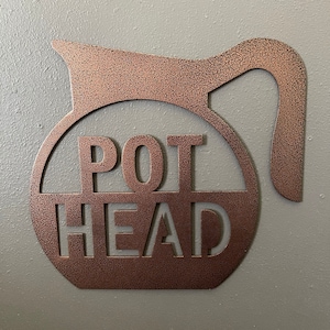 May include: A copper-colored metal wall art piece in the shape of a coffee pot. The words "POT HEAD" are cut out of the metal within the pot's body. The art is mounted on a gray wall.