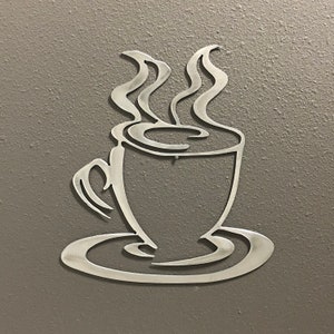 May include: A metal wall decor of a coffee cup with steam rising from it. The cup is on a saucer.