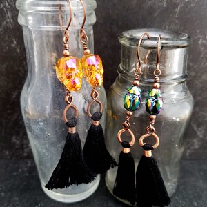 May include: Two pairs of earrings with black tassels. One pair features iridescent orange skull beads and the other pair features iridescent green scarab beetle beads.