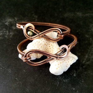 Pode incluir: Duas pulseiras de fio de cobre em forma de símbolo de infinito com um detalhe de pérola verde. As pulseiras estão empilhadas uma em cima da outra e estão apoiadas em uma superfície branca semelhante a coral.