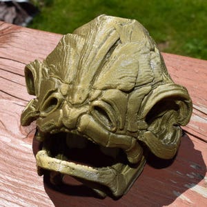 May include: A 3D printed, gold-colored, detailed creature head with a wide open mouth. The creature has a textured, scaly appearance and large, round eyes.