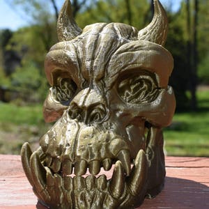 May include: A gold-colored 3D printed skull with horns and sharp teeth. The skull is detailed with textured ridges and appears to be made of a plastic material.