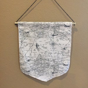 May include: A white canvas banner with a black and white vintage world map print. The map features airplane routes and compass directions. The banner is hung from a wooden dowel rod.