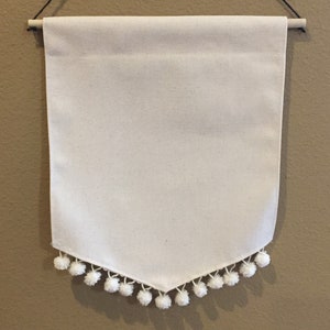 May include: A white canvas banner with a white pom-pom trim. The banner is hanging from a wooden dowel rod with black string.