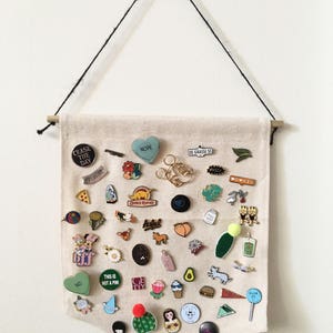 May include: A beige fabric banner with a wooden dowel at the top and bottom. The banner is covered with a variety of enamel pins, including a heart-shaped pin that says "NOPE", a pin that says "CEASE THE DAY", and a pin that says "DE GRASSI ST".
