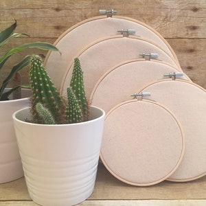 May include: Five wooden embroidery hoops of varying sizes, with natural colored fabric stretched across them. The hoops are arranged in a semi-circle, with the largest hoop at the top and the smallest hoop at the bottom. The hoops are sitting on a wooden surface.
