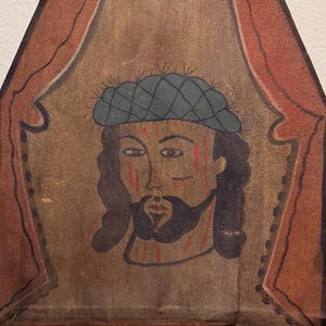 May include: Hand-painted religious icon featuring a portrait of a man with a beard, wearing a blue cap and a crown of thorns. The artwork is framed by a red curtain design. The icon is on a weathered, aged material.