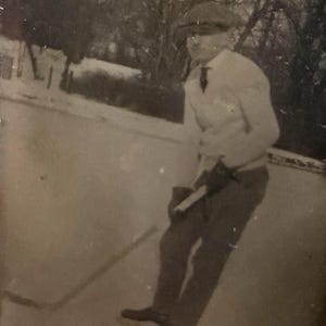 May include: Vintage black and white photograph of a person playing ice hockey. The person is wearing a cap, tie, and gloves, holding a hockey stick. The background shows trees and a snowy landscape.