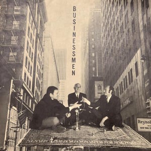 May include: Black and white photograph of three people in suits sitting on a rug in a city setting. The word "Businessmen" is vertically printed in the image.