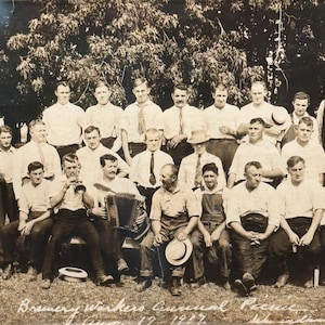 May include: A black and white vintage photograph of a large group of men posing outdoors in front of trees. The men are dressed in light-colored shirts and trousers, with some wearing hats. One man holds a trumpet and another plays an accordion.