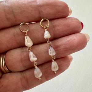 May include: Gold-tone dangle earrings featuring three off-white, teardrop-shaped beads. The beads are strung together with gold wire and connected to a gold hoop. The earrings are displayed against a neutral background.