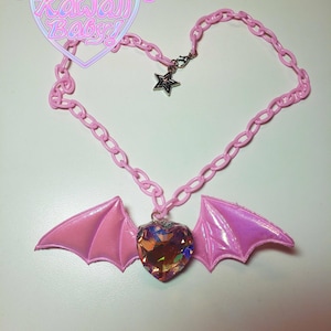 May include: A pink chain necklace with a heart-shaped pendant and bat wings. The pendant is a multi-colored crystal. A silver star charm hangs near the clasp. The text "Pastel Kawaii Baby!" is in a heart-shaped frame.
