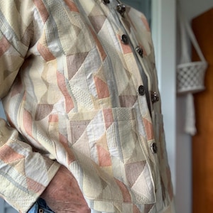 May include: A beige, peach, and brown patchwork jacket with a geometric design. The jacket has a mandarin collar, button closures, and two front pockets. The jacket is worn with a white shirt and blue jeans.