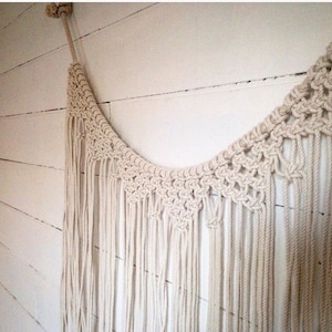 May include: A white macrame wall hanging with a curved shape and long fringe. The hanging is made of natural cotton rope and has a bohemian style.