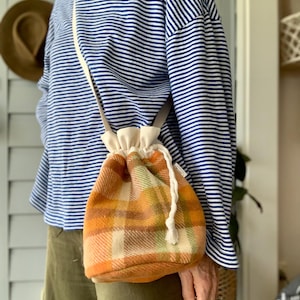 May include: A plaid bucket bag in orange, brown, and green. It has a cream drawstring closure and strap. The background shows a blue and white striped top and olive green trousers. The bag's design is reminiscent of a traditional Scottish tartan.