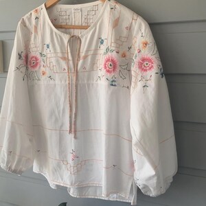May include: A white long-sleeve blouse featuring a round neckline with a tie closure, balloon sleeves with elastic cuffs, and intricate floral embroidery in shades of pink, blue, and orange, accented by geometric line patterns across the chest.