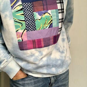 May include: A light blue sweatshirt with a tie-dye pattern and a large patchwork pocket on the front. The pocket is made of various fabrics in shades of purple, blue, green, and white.