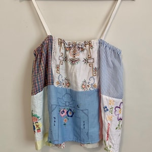May include: A sleeveless patchwork camisole top featuring a variety of fabric panels including white linen with colourful floral embroidery, blue and white striped cotton, and a blue panel with a central floral design, all held by thin white straps.