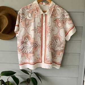 May include: White short-sleeved shirt with a repeating pattern in red and black. The pattern features stylized birds and geometric shapes. The shirt is hanging on a wooden hanger.