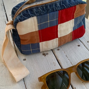 May include: A rectangular zipped pouch made from blue denim and a patchwork of red, white, tan, and blue plaid fabric squares. The pouch features a beige herringbone wrist strap and is positioned next to a pair of amber-coloured sunglasses.