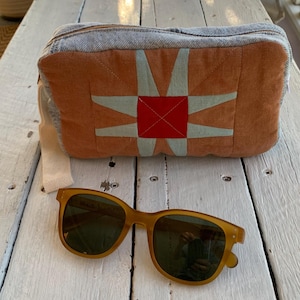 May include: A rectangular zipped pouch made of light blue denim and orange fabric with a central red square and light blue star-shaped appliqué. A pair of amber-coloured sunglasses with dark lenses is positioned in front of the pouch.