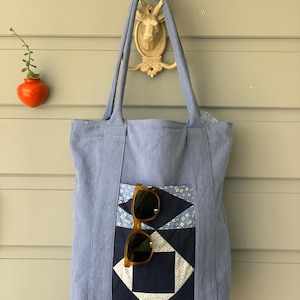 May include: A light blue tote bag with a patchwork design in shades of blue and white. The bag has a large pocket on the front with a pair of brown sunglasses inside.