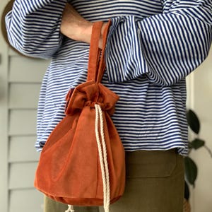 May include: A burnt orange velvet drawstring bag with a cream rope closure. The bag is held by a person wearing a blue and white striped top and olive green trousers. The bag has a soft, plush texture.