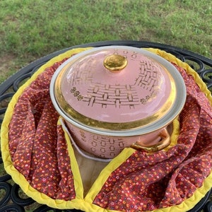 May include: A pink and gold ceramic casserole dish with a woven pattern sits on a red and yellow floral fabric. The dish has a lid and a gold handle.
