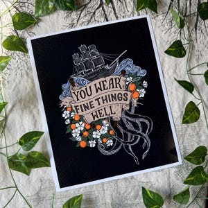 以下が含まれることがあります： 黒色の背景に帆船、タコの触手、オレンジの花を描いたアートプリント。「YOU WEAR FINE THINGS WELL」というテキストがバナーに表示されています。緑の葉が画像を囲み、装飾的な効果を生み出しています。