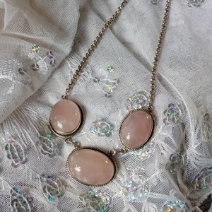 Pu&ograve; includere: Una collana a catena d'argento con tre pietre ovali di quarzo rosa. La collana &egrave; su uno sfondo di pizzo bianco.