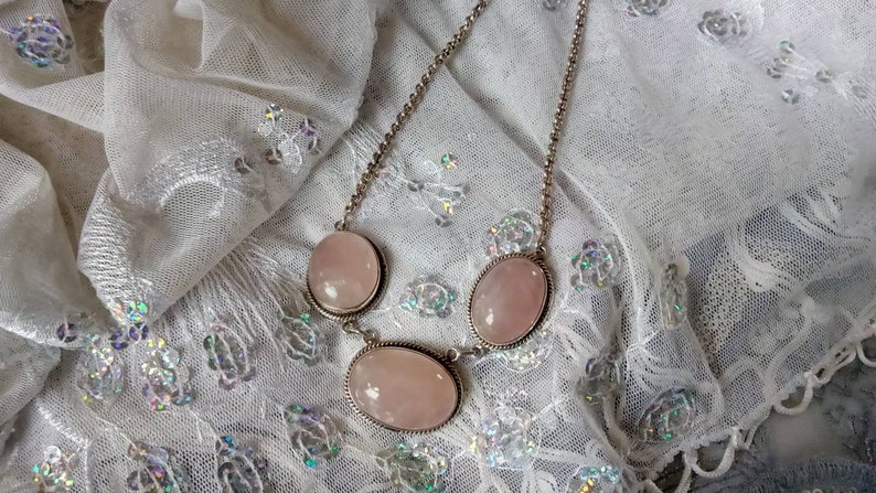 Pu&ograve; includere: Una collana a catena d'argento con tre pietre ovali di quarzo rosa. La collana &egrave; su uno sfondo di pizzo bianco.