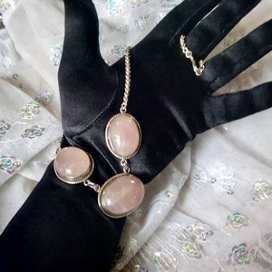 Pu&ograve; includere: Un braccialetto a catena d'argento con tre pietre ovali di quarzo rosa. Il braccialetto &egrave; su un guanto nero.