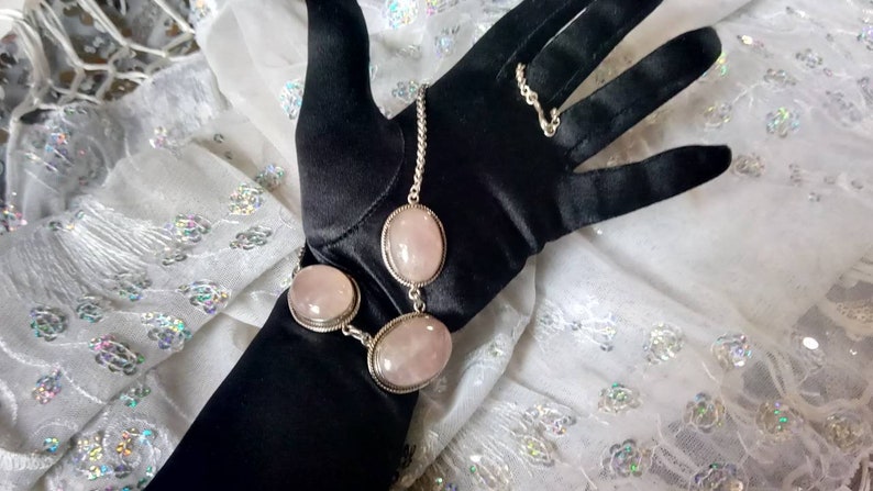 Pu&ograve; includere: Un braccialetto a catena d'argento con tre pietre ovali di quarzo rosa. Il braccialetto &egrave; su un guanto nero.