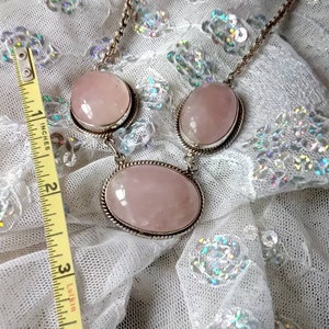 Pu&ograve; includere: Una collana d'argento con tre grandi pietre ovali di quarzo rosa pallido. La collana &egrave; su un tessuto bianco scintillante.