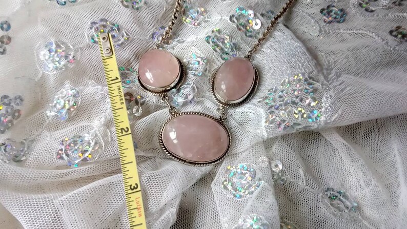 Pu&ograve; includere: Una collana d'argento con tre grandi pietre ovali di quarzo rosa pallido. La collana &egrave; su un tessuto bianco scintillante.