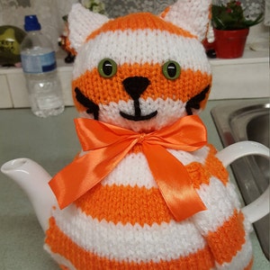 May include: A white ceramic teapot with a knitted cat tea cosy. The cosy is striped orange and white, with green button eyes, a black nose and whiskers, and an orange ribbon bow. The cat cosy is designed to keep the tea warm.