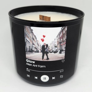 May include: Black candle jar with a white label featuring a photo of a couple kissing in front of a city street with red heart balloons. The label also includes the song title "Glow" by "Alien Ant Farm" and a music player icon with play, pause, and skip buttons.