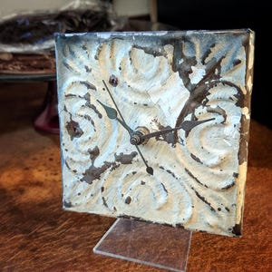 May include: Square, distressed metal clock with a cream-colored face and ornate embossed design. The clock has black hands and a clear plastic stand. The metal frame has a dark, weathered appearance, adding to its vintage charm.