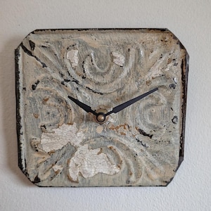May include: Square wall clock with a distressed, vintage look. The clock face is a light blue-gray with a floral pattern and black hands. The clock's frame is dark brown with a slightly weathered appearance.