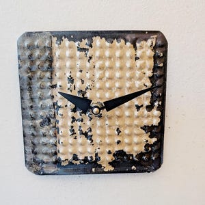 May include: Square wall clock with a textured, distressed metal face. The clock face is primarily beige with black and gray accents. Black clock hands point to the time. The clock has a vintage, industrial aesthetic.