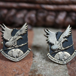 Puede incluir: Par de gemelos Air Defense Command vintage. Cada gemelo presenta un diseño de escudo con un emblema de águila detallado, un rayo y las palabras "Air Defense Command". Los gemelos están hechos de metal plateado.