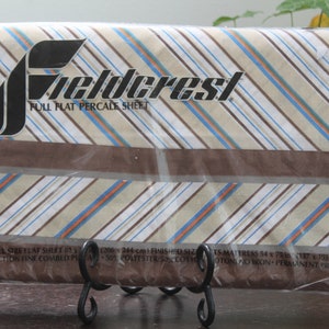 May include: A full flat percale sheet in a plastic package. The sheet has a striped pattern in beige, brown, blue, and orange. The brand name "Fieldcrest" is printed in black. The sheet is designed for a full-size mattress.