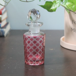 Puede incluir: Un pequeño frasco de perfume de vidrio transparente con un tapón de cristal facetado. El frasco tiene un exterior rosa con un diseño de diamantes. El frasco está sobre una superficie de madera oscura, con una pila de libros y una planta en maceta en el fondo.