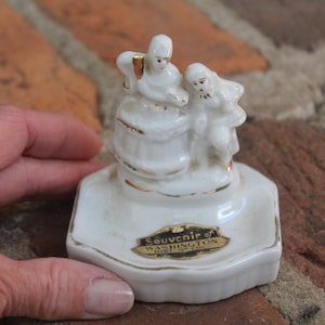 May include: A white ceramic souvenir figurine featuring two figures, with gold trim accents. The base is a hexagonal shape with the text "Souvenir of Washington D.C." in gold lettering. The figurine is held by a hand.