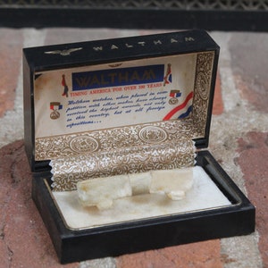 May include: A vintage black watch box with gold-colored interior lining and a white velvet insert. The box lid features the name "WALTHAM" and text that reads "TIMING AMERICA FOR OVER 100 YEARS".