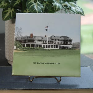 May include: Square ceramic tile featuring a color image of a large building with a green lawn and a flagpole. The text "THE ROCKAWAY HUNTING CLUB" is printed at the bottom. The tile is displayed on a gold-colored metal stand.