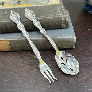 Può includere: Una forchetta e un cucchiaio da portata d'argento con dettagli floreali ornati. La forchetta ha tre rebbi e il cucchiaio ha una coppetta forata. I manici presentano un design decorativo. Le posate sono esposte su una superficie di legno scuro accanto a libri d'epoca.