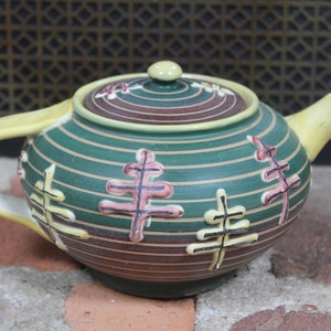 May include: A ceramic teapot with a green and brown striped pattern, adorned with stylized tree designs in pink and yellow. The teapot features a yellow handle, spout, and lid.